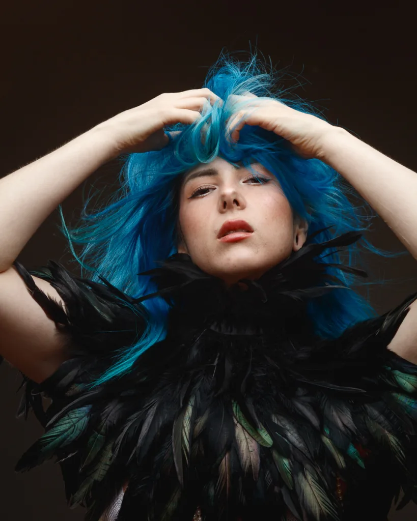 Person with blue hair wears feathered outfit.