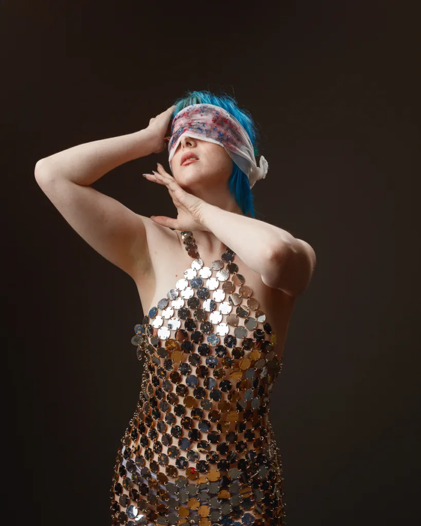 Person with blue hair and blindfold in shiny dress.