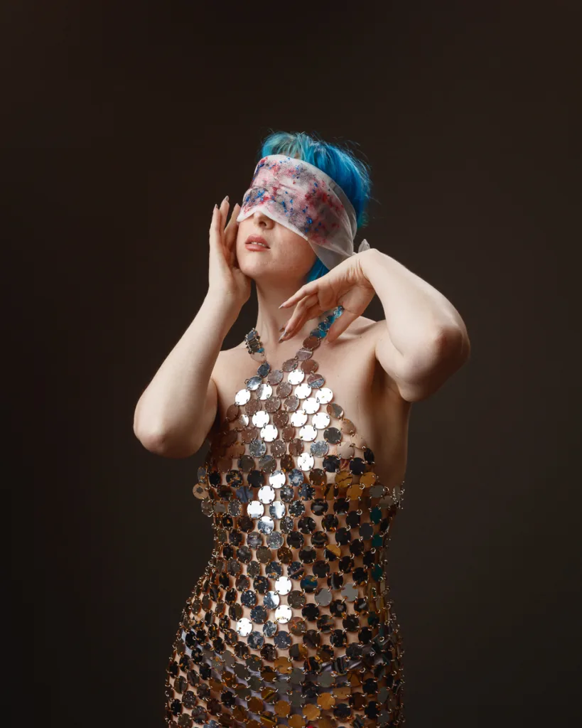 Person with blue hair, metallic dress, blindfolded.