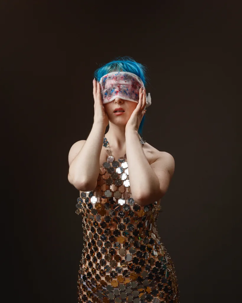 Woman with blue hair wearing a sequined dress, blindfolded.