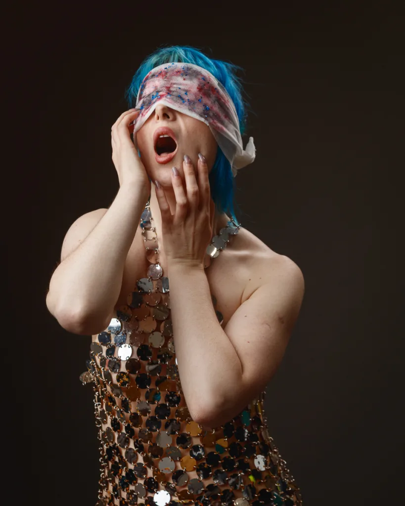 Person with blue hair and decorated dress blindfolded.