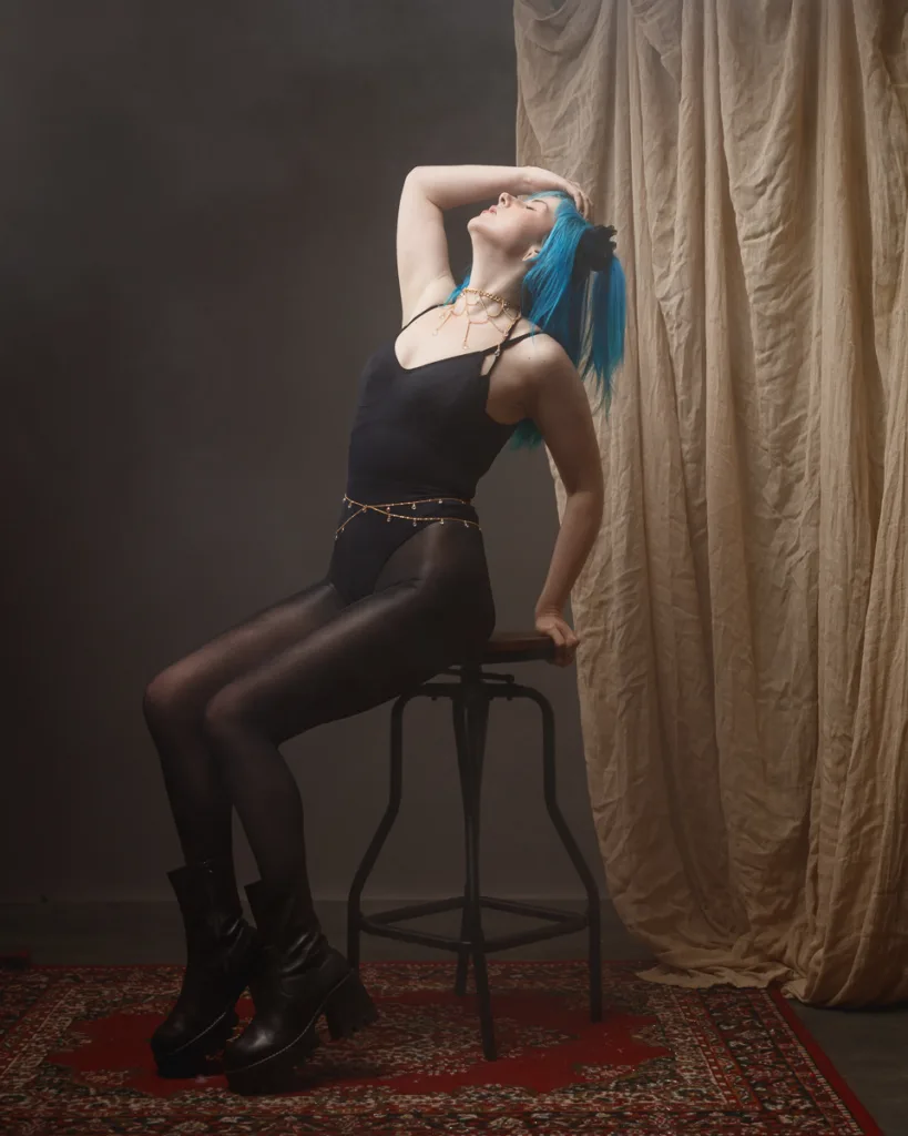 Woman with blue hair posing on stool indoors.