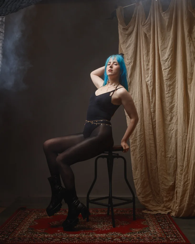 Person with blue hair posing on stool, dramatic lighting.