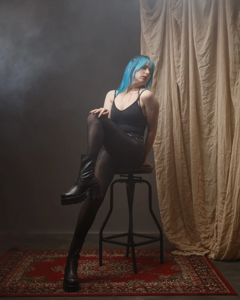 Fashionable person with blue hair on stool in studio.