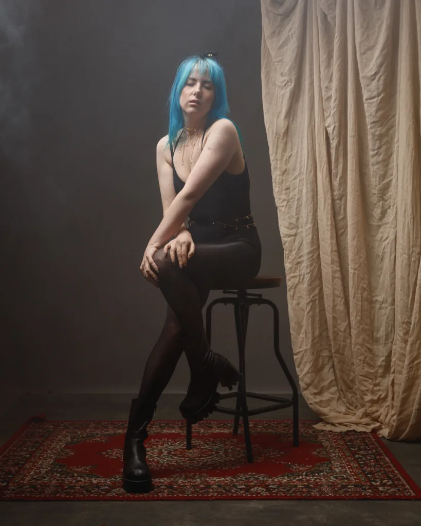 Person with blue hair sitting on a stool indoors.