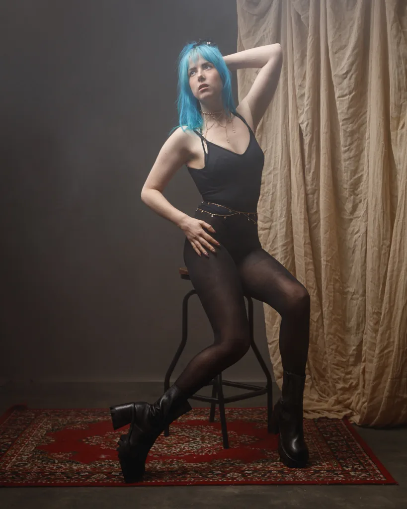 Person with blue hair posing on stool, studio backdrop.