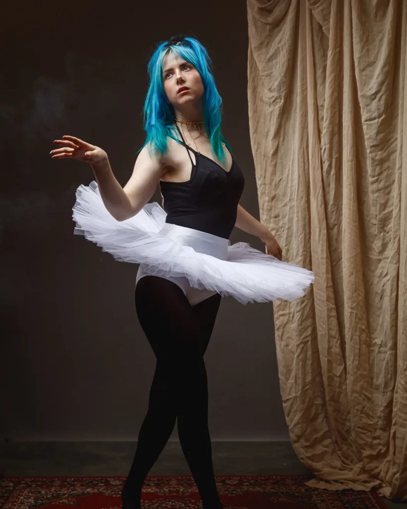 Dancer with blue hair wearing white tutu, posing gracefully.
