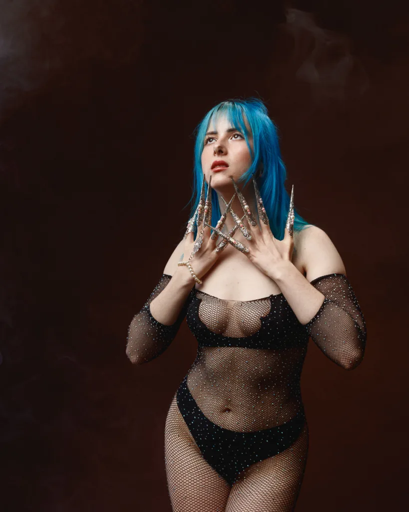 Person with blue hair, ornate long nails, and fishnet outfit.