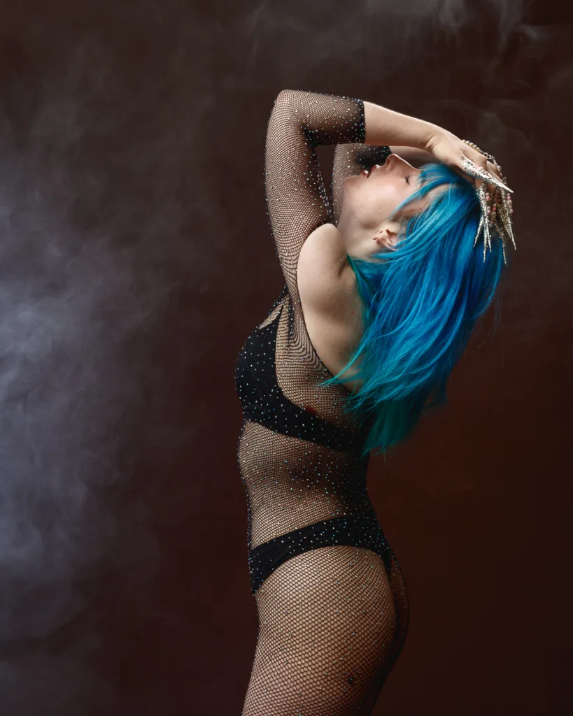 Person with blue hair posing dramatically in mesh outfit.