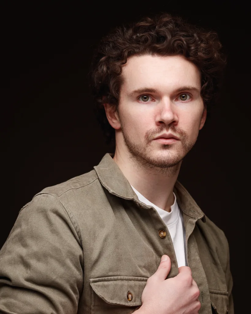 Man in khaki jacket posing against dark background.