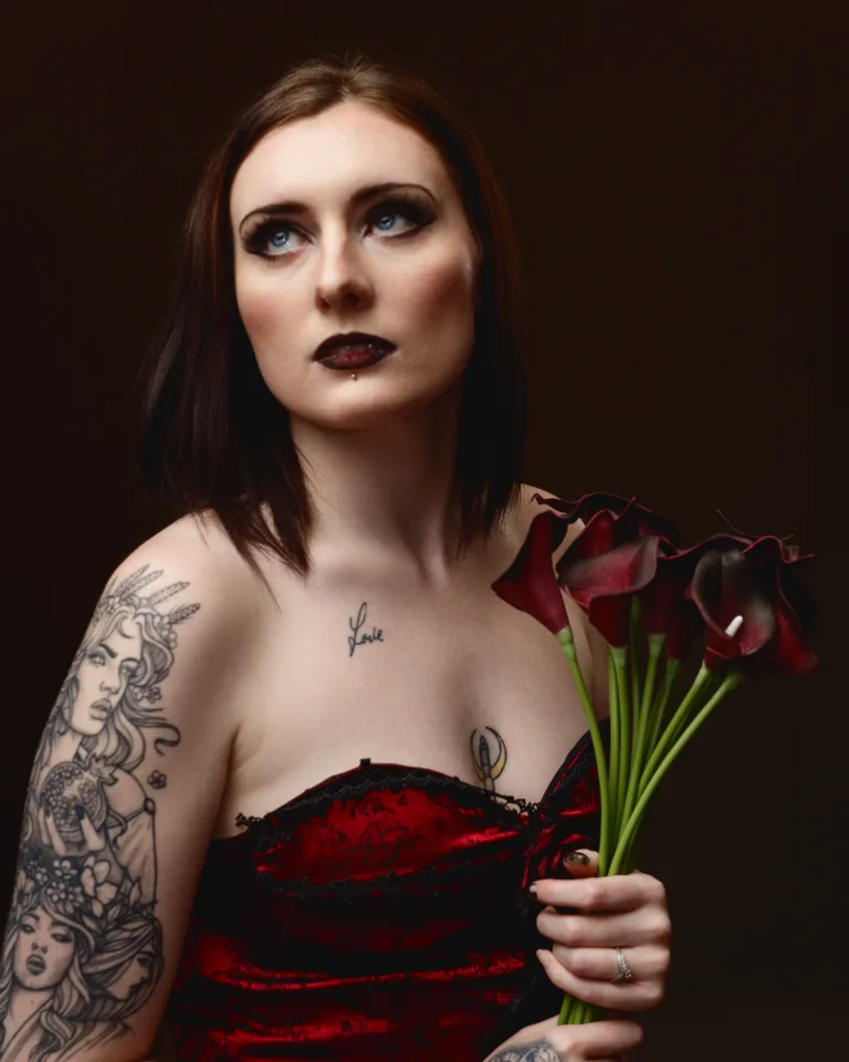 Tattooed woman in red dress holding dark flowers.