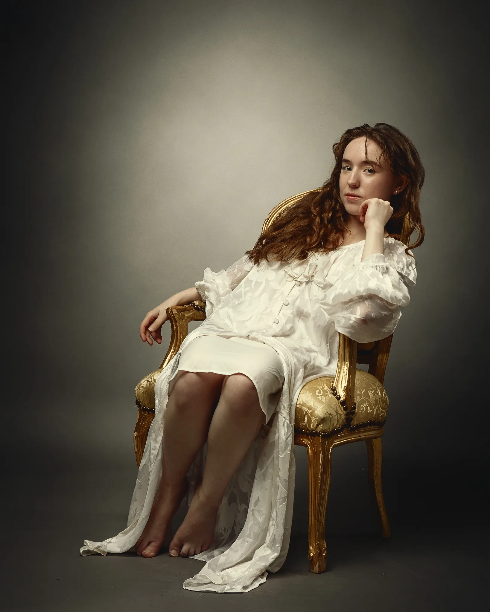 A young woman with long brown wavy hair sits on a gilt wooden chair wearing a white flowing dress, gazing toward the camera with her hand raised to her face.