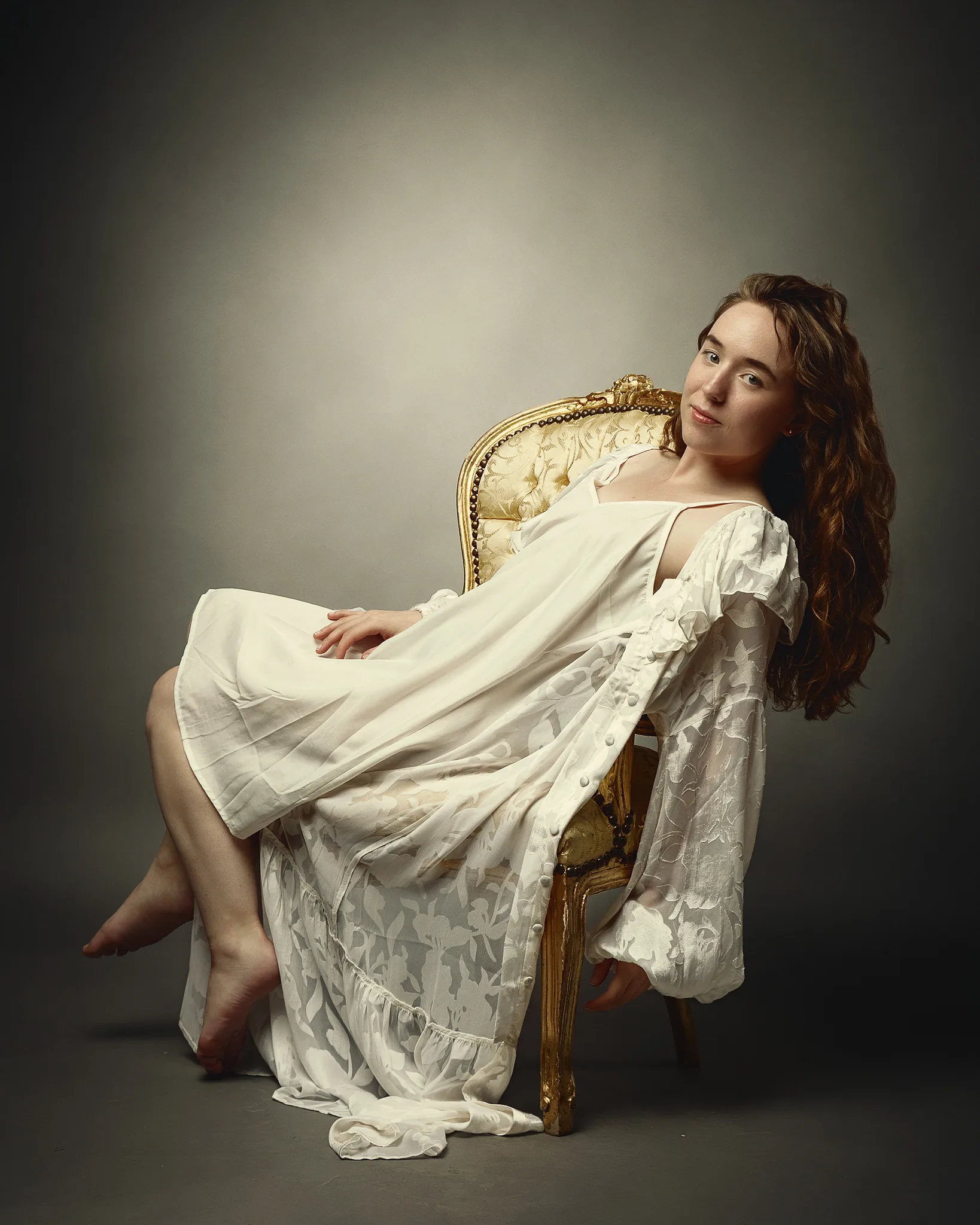A woman in a cream linen dress reclines on a gilded Rococo chair against a neutral background, looking toward the camera.