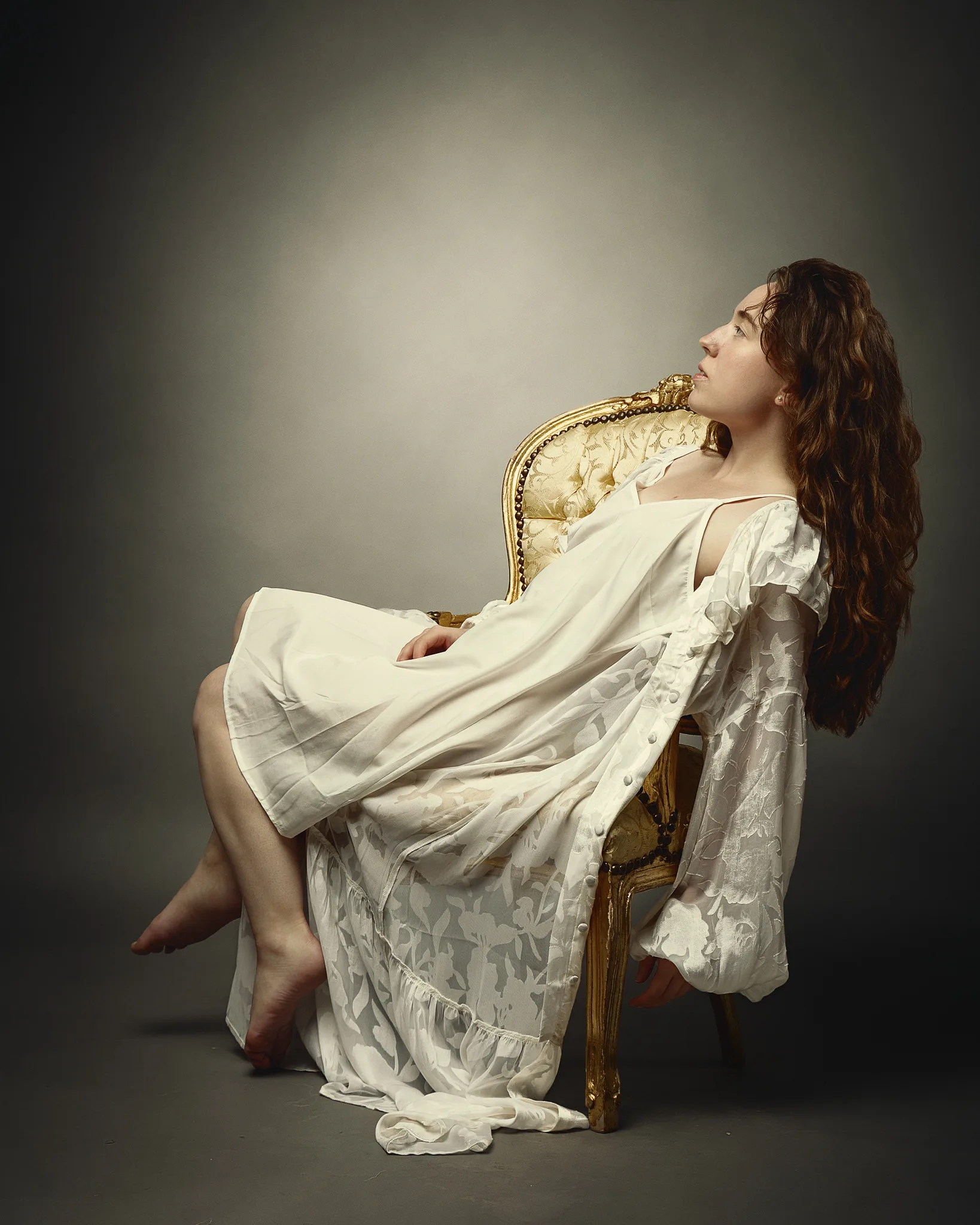 A woman with long brown hair sits on an ornate gilt chair, draped in flowing white fabric, gazing upward against a neutral background.