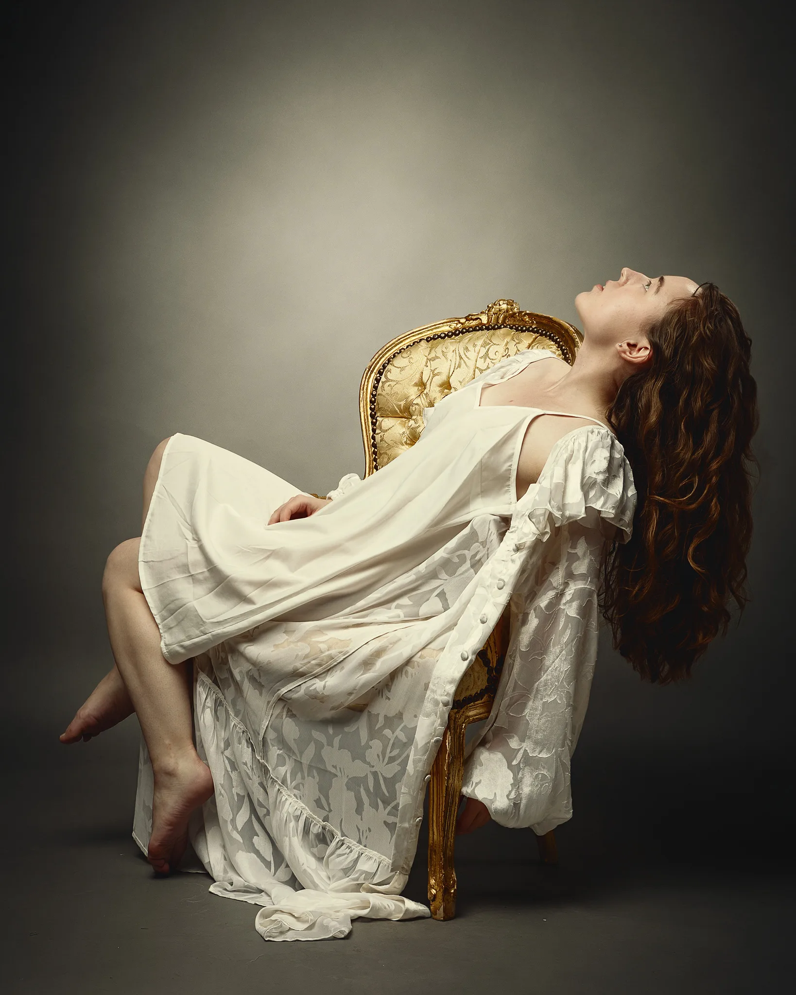 A woman in a white draped garment reclines on an ornate gold chair against a neutral background, head tilted back.