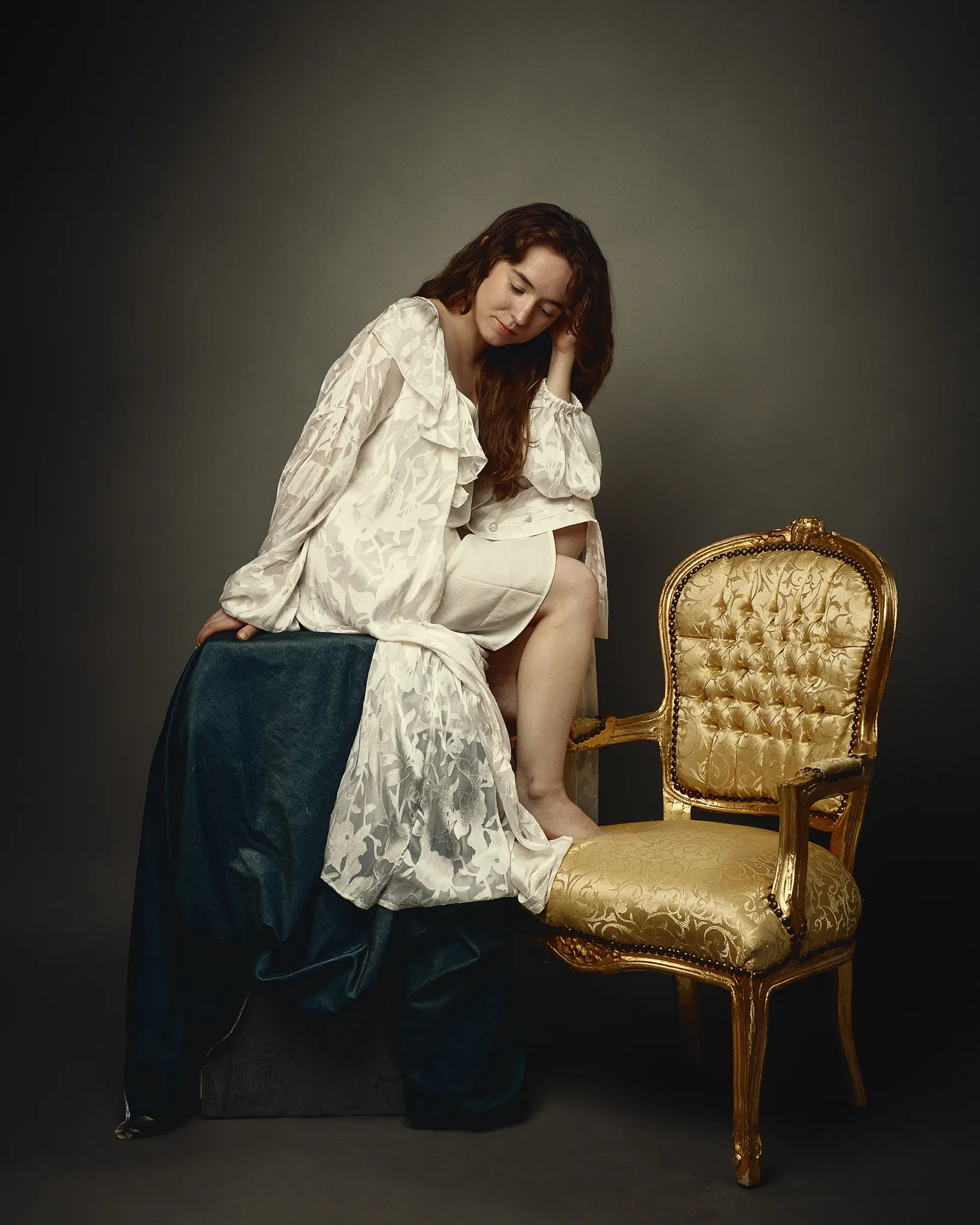 A woman in a white patterned blouse sits on a dark blue fabric, leaning toward an ornate gold chair with a contemplative expression.