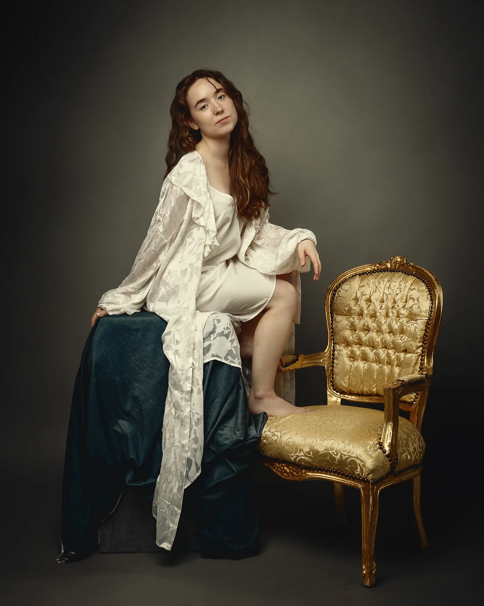A woman with long wavy brown hair sits beside a gold ornate chair, wearing cream and teal clothing against a neutral background.