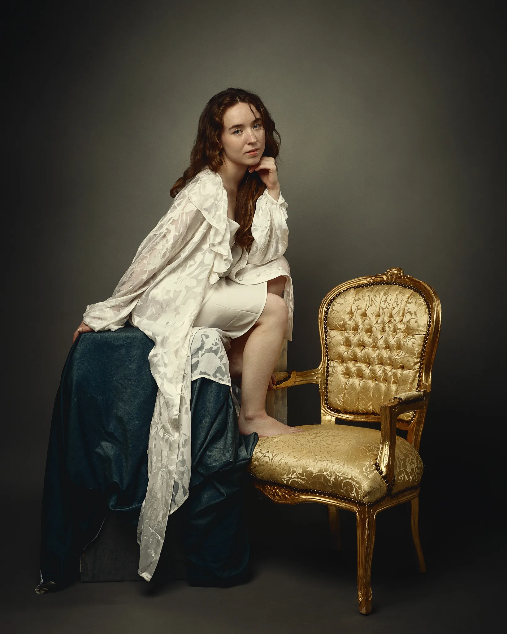 A woman sits on a gilded chair against a neutral background, wearing a white ruffled blouse and dark skirt.
