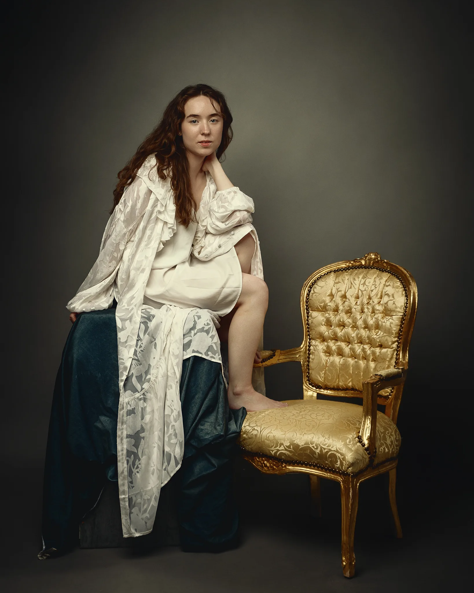 A young woman with long reddish-brown hair sits posed next to an ornate gilt chair, wearing a cream blouse and dark skirt with a white shawl draped over her shoulder, against a neutral grey background.