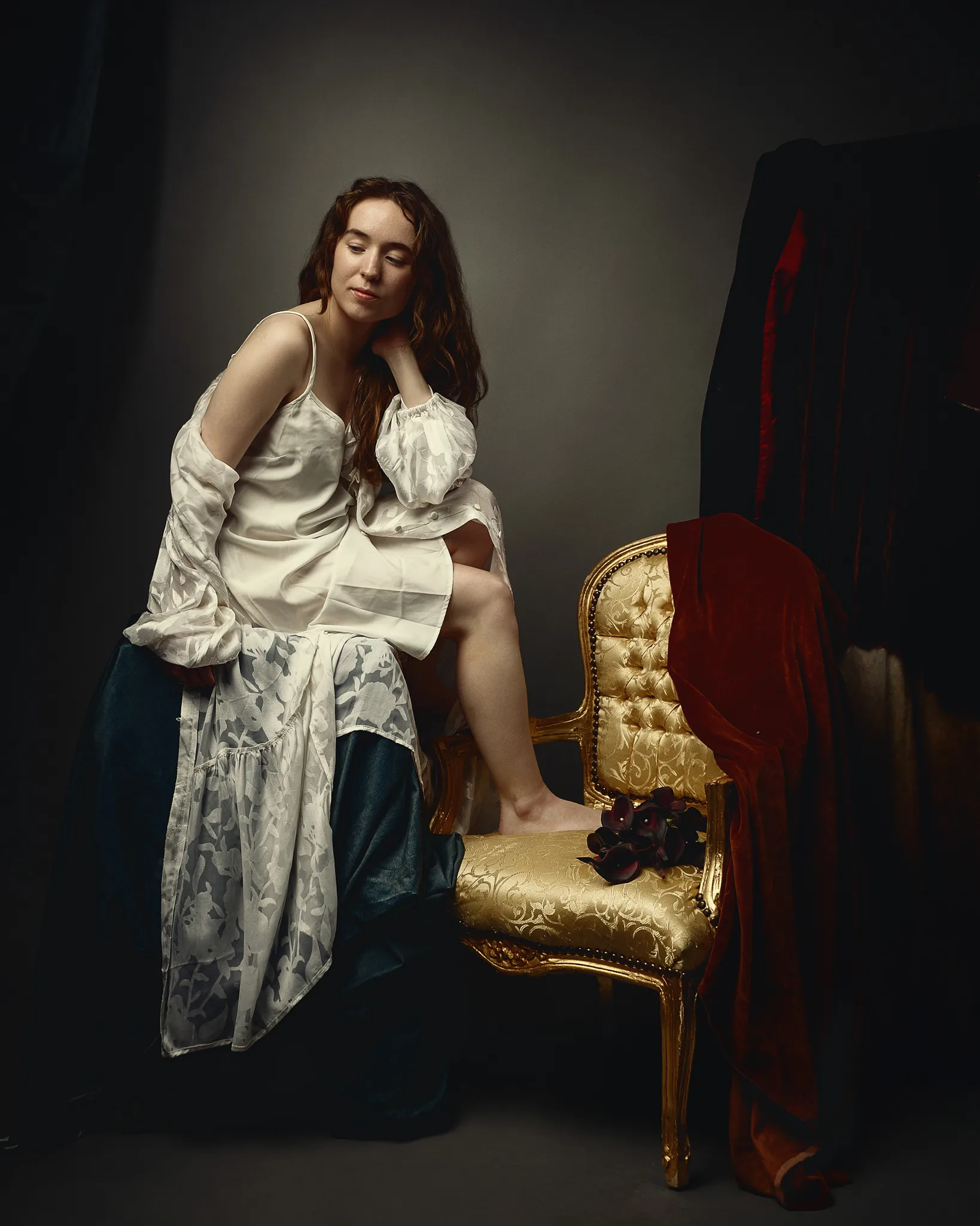 Woman in white draped clothing sitting on ornate gold chair with red velvet draping in dark studio setting.