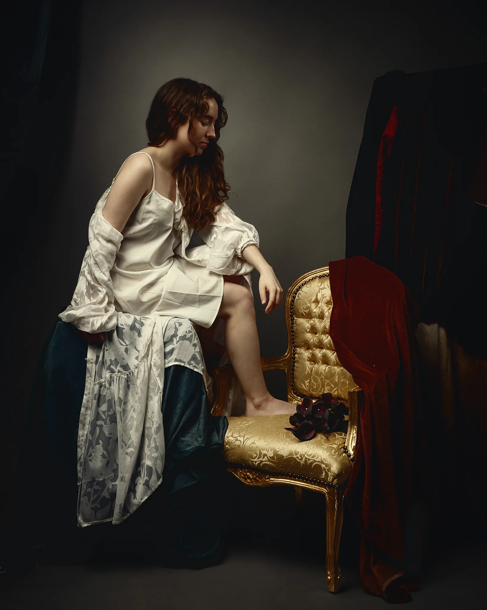 A woman in a white silk robe sits on a gold-trimmed chair with red velvet upholstery against a dark background.