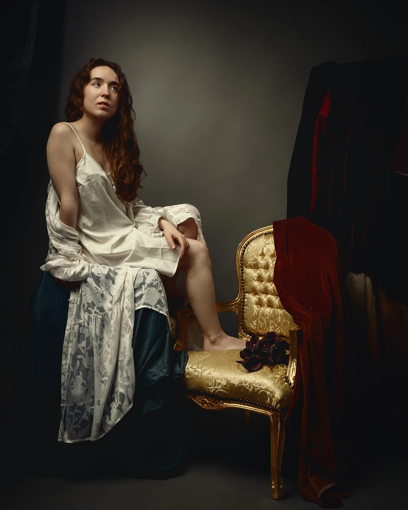 A woman in a white slip dress stands beside an ornate gold chair draped with red fabric against a dark background.