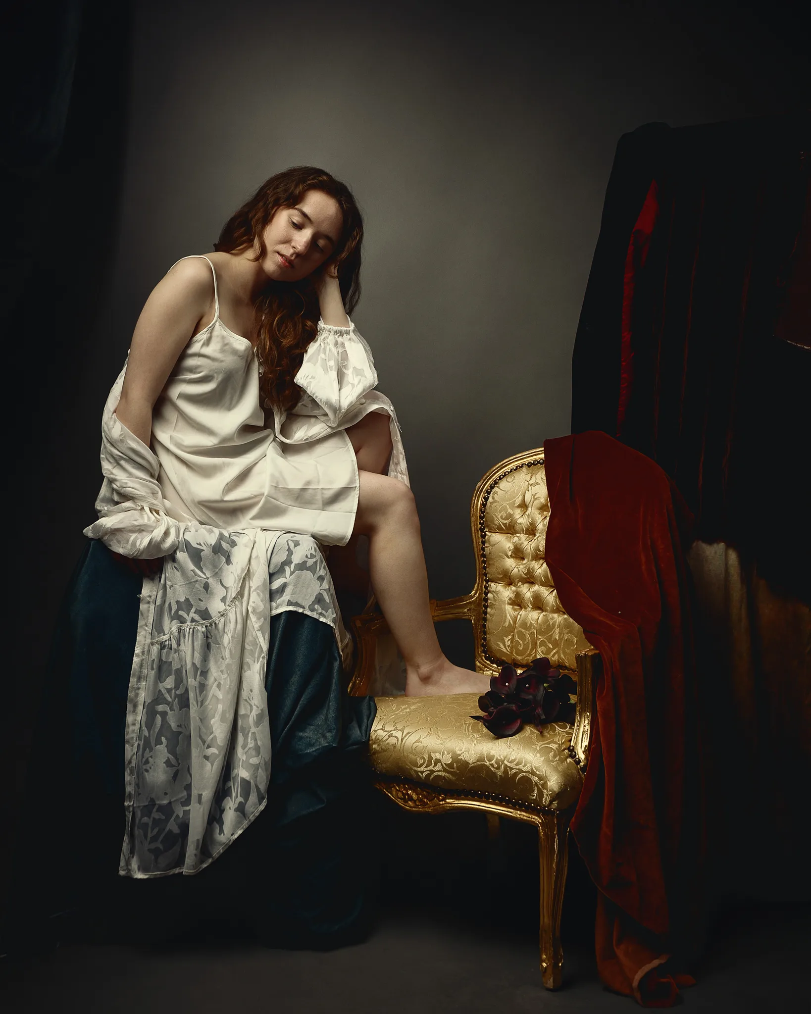 Woman in white chemise and dark skirt sitting on ornate gilt chair draped with red fabric, head tilted pensively.