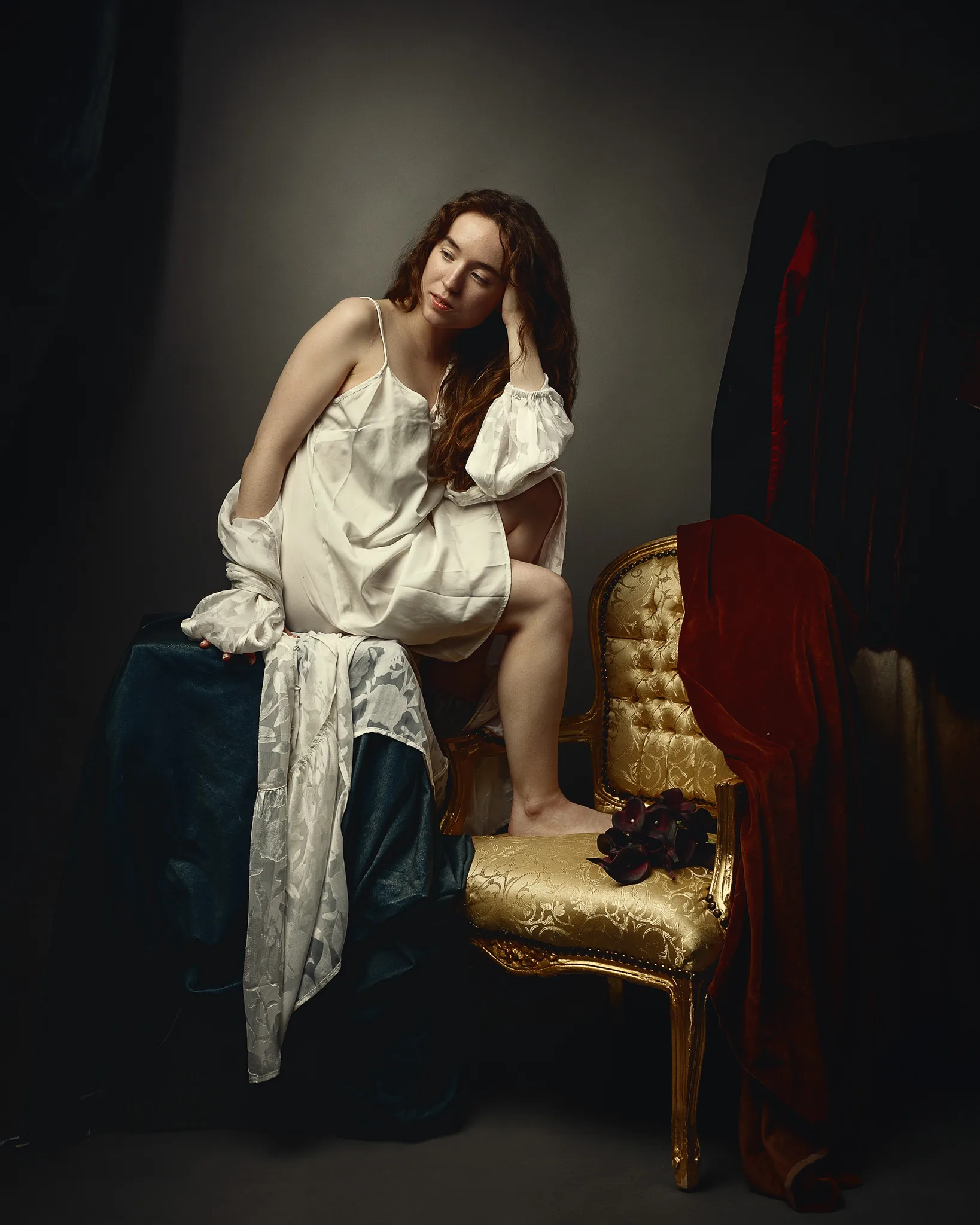 Woman in white chemise seated on ornate gold chair with red velvet drape, hand to face, dark background.
