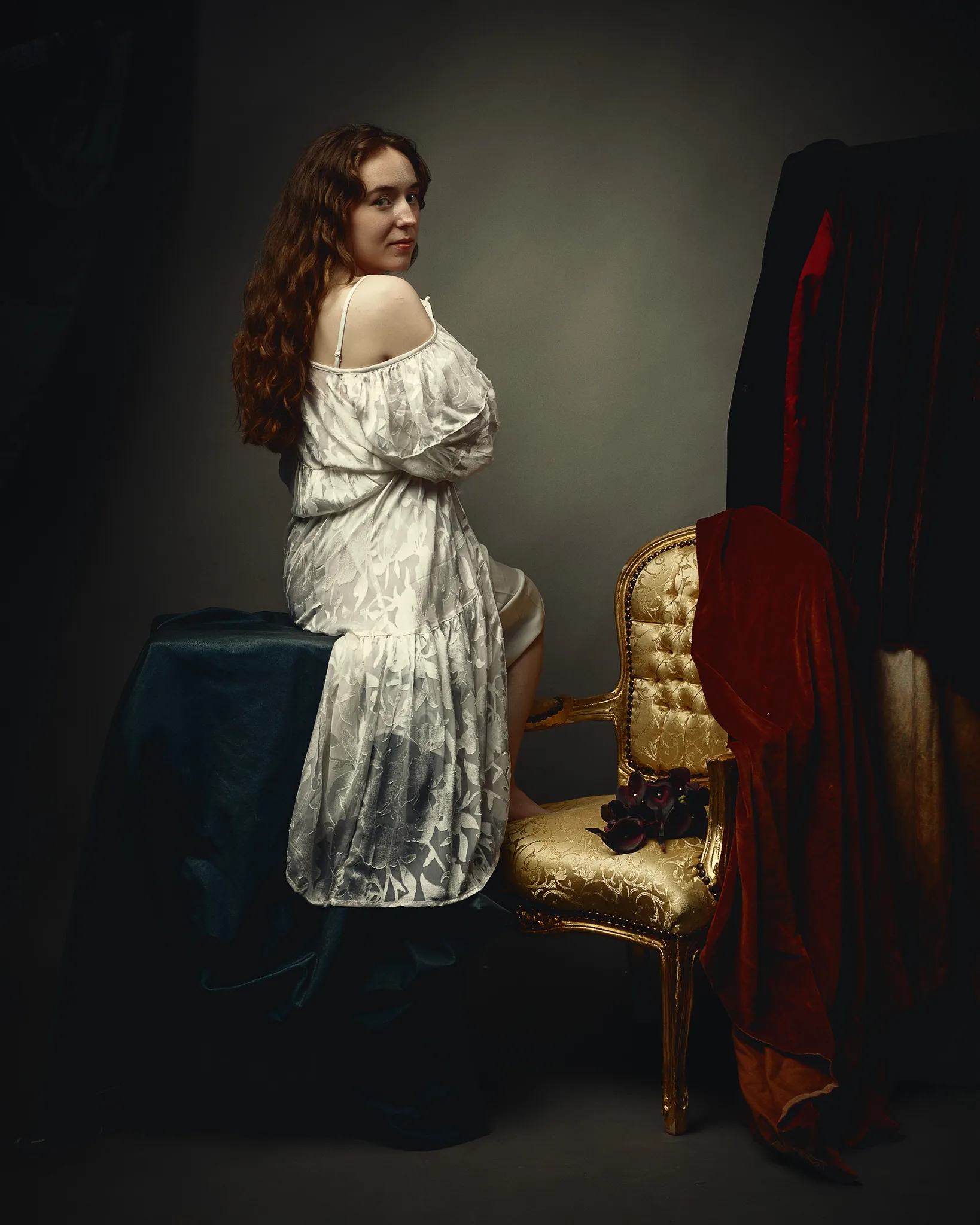 A woman with long reddish-brown hair wearing a white off-shoulder dress stands beside a gold chair draped with red fabric.