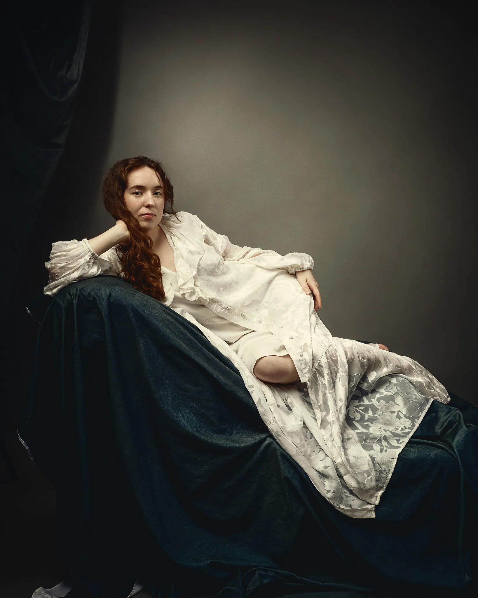 A woman with long reddish-brown hair sits on a dark blue velvet chair against a grey background, wearing a cream blouse and gazing to the side.
