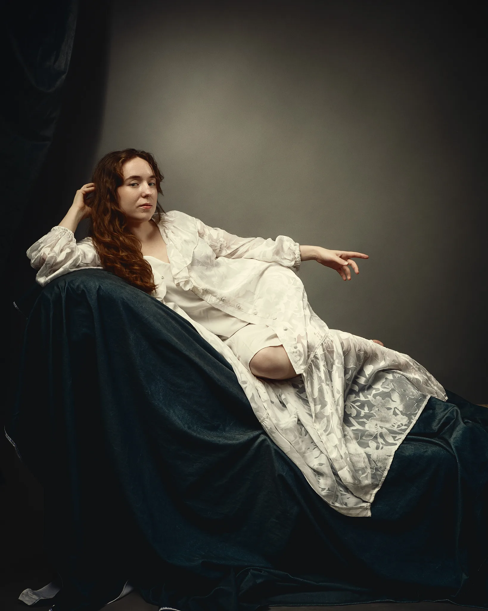 A woman with long reddish-brown hair reclines on dark fabric, wearing a cream-coloured dress, gazing to the side thoughtfully.