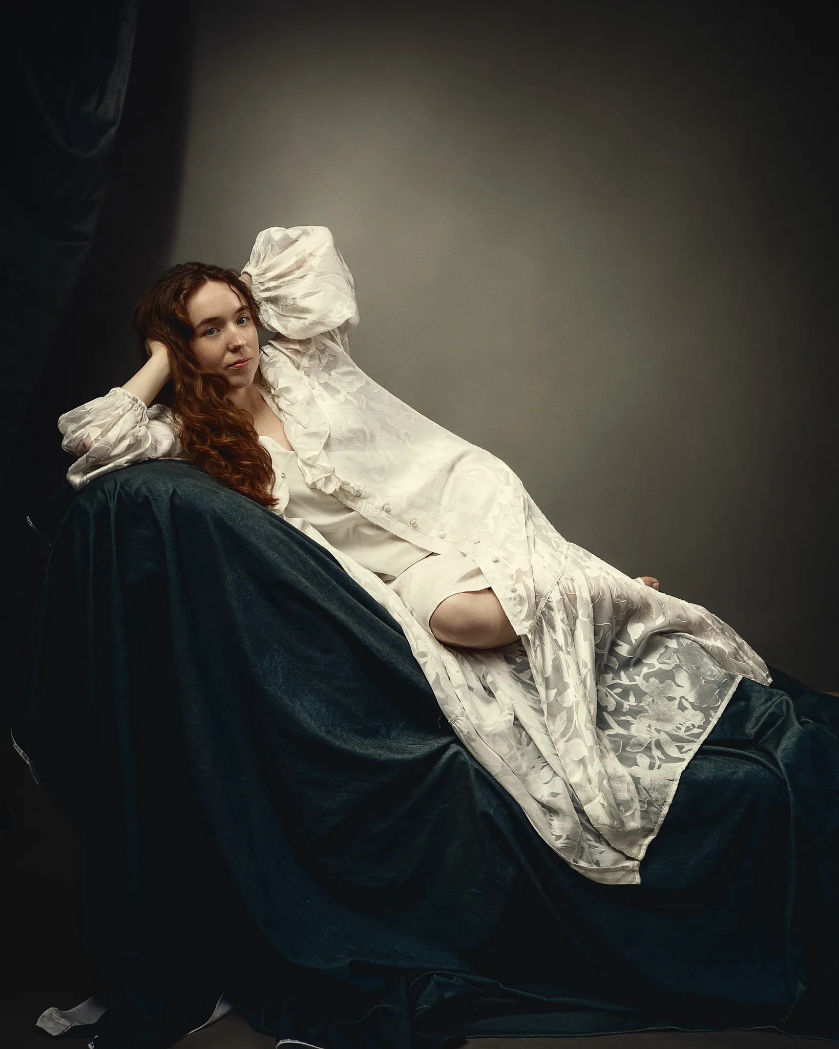 A woman with long reddish-brown hair sits against a dark background, wearing a cream-colored robe over a dark teal dress, gazing thoughtfully to the side.