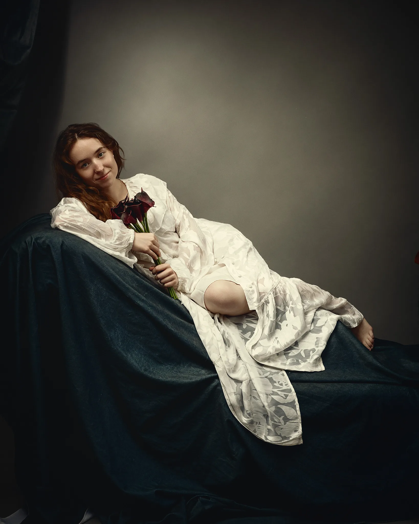 A young woman in a white dress sits on dark fabric, holding dark red flowers, gazing toward the camera with a slight smile.