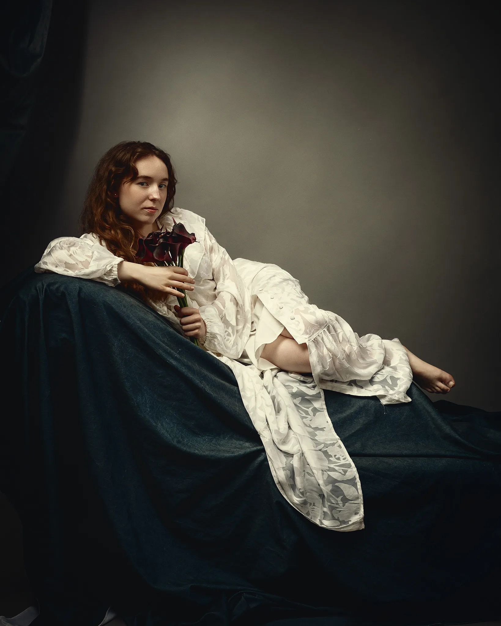 A young woman with reddish-brown hair sits on a blue fabric, wearing a white ruffled dress and holding dark red flowers. Neutral background.