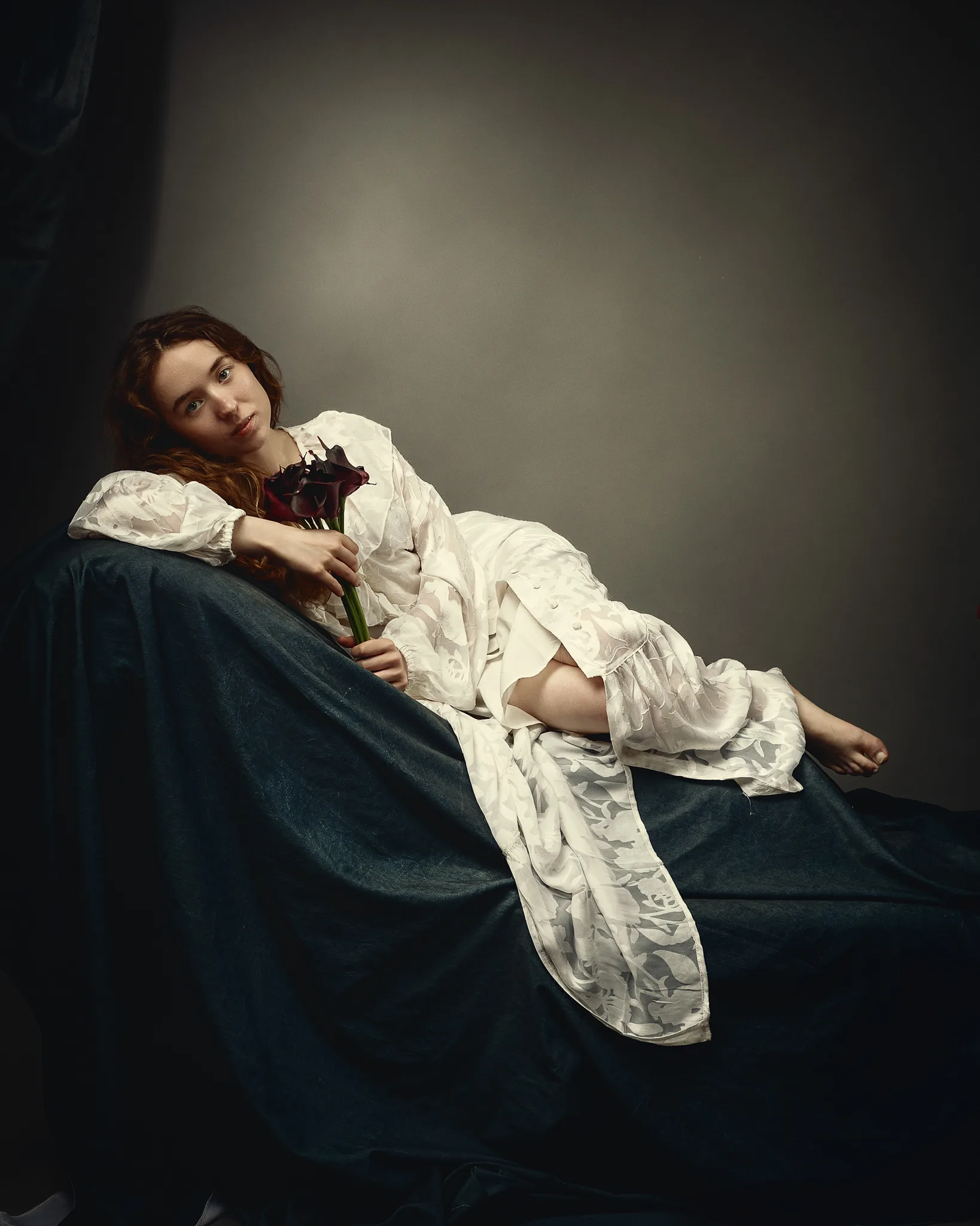 A woman in a white chemise reclines on dark fabric, holding a dark red flower, gazing toward the camera against a neutral background.