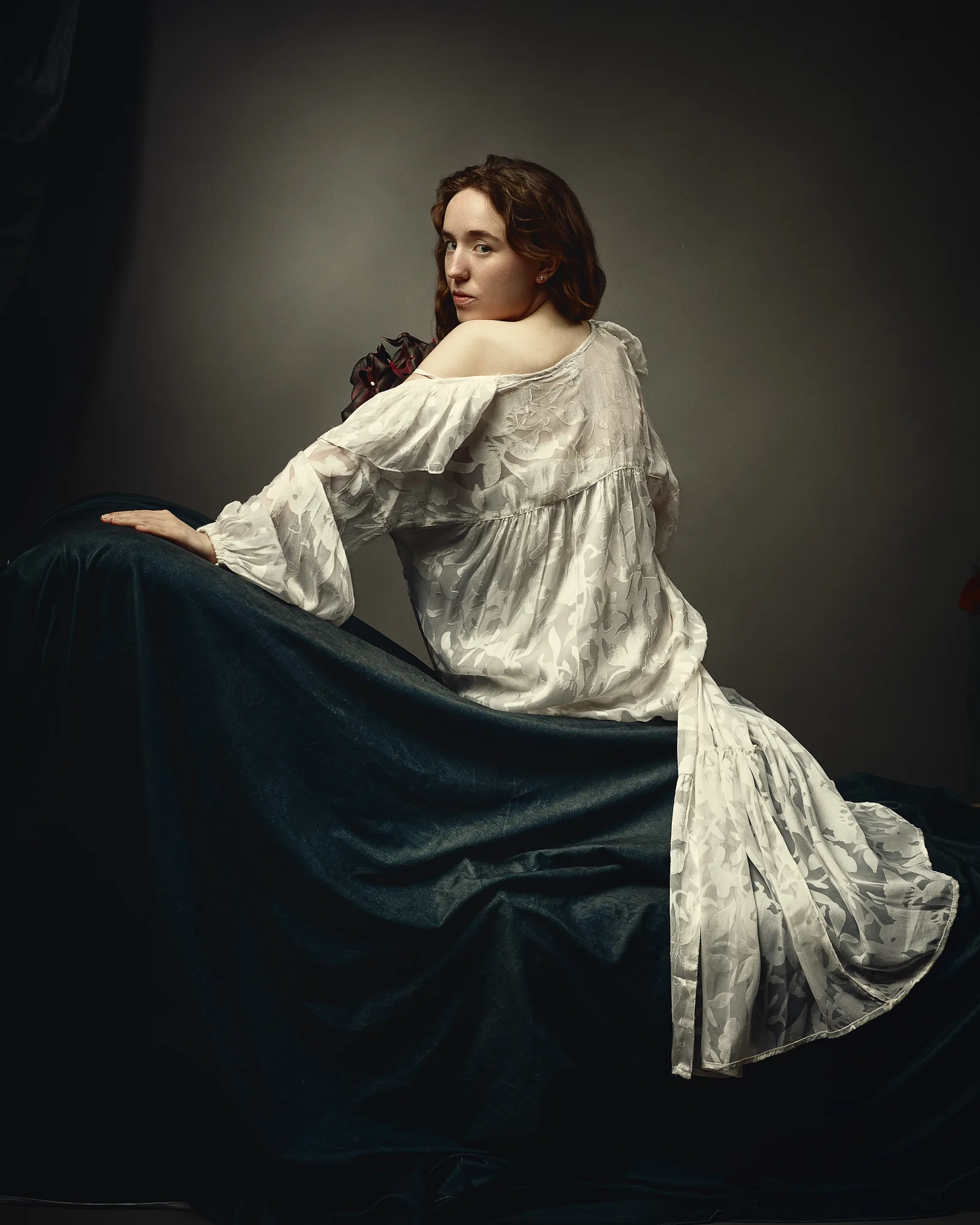 A young woman in a white flowing gown reclines on dark blue fabric, looking over her shoulder at the viewer against a neutral background.