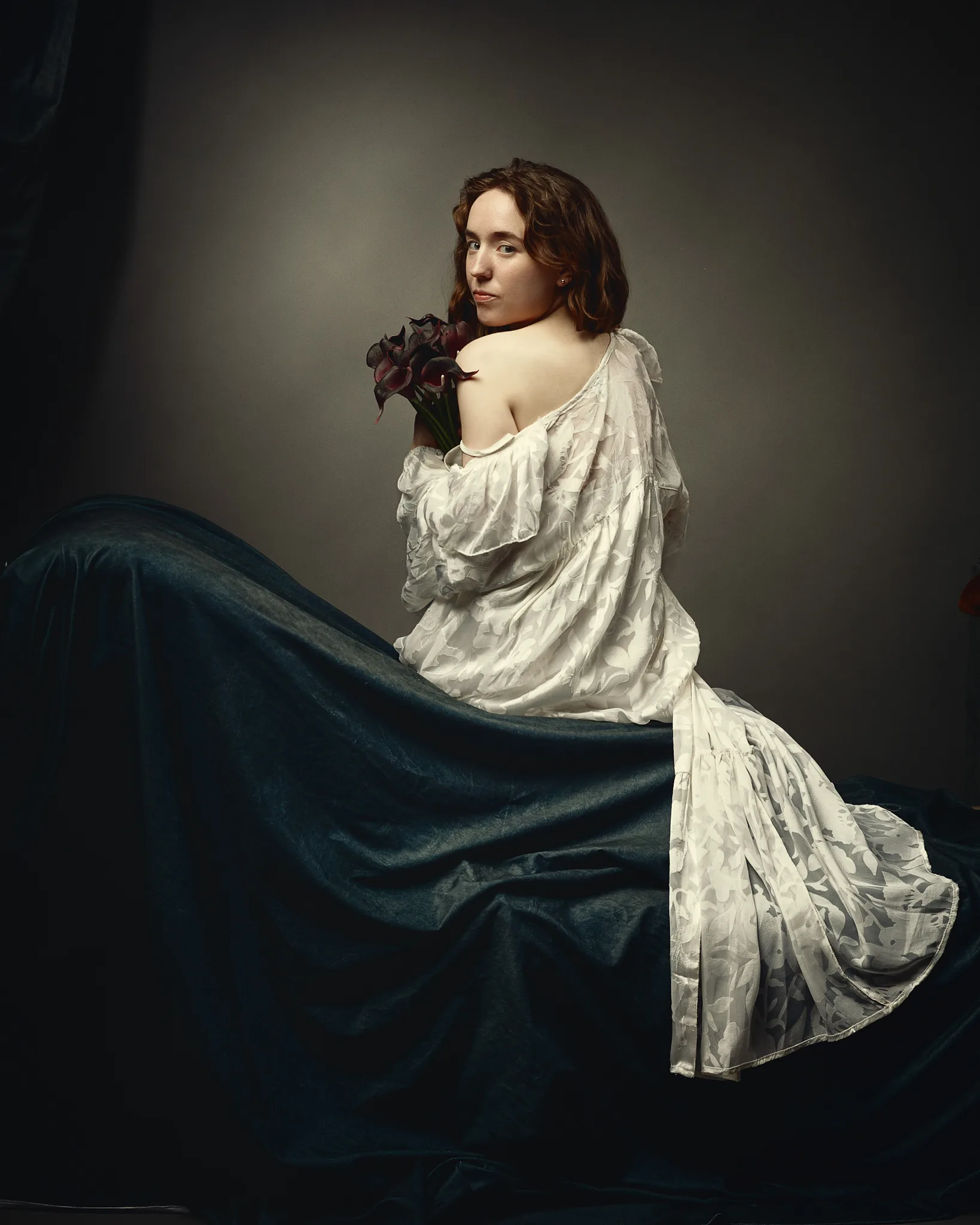 A woman with brown wavy hair holds dark red flowers over her shoulder, wearing a white draped garment and dark blue fabric.
