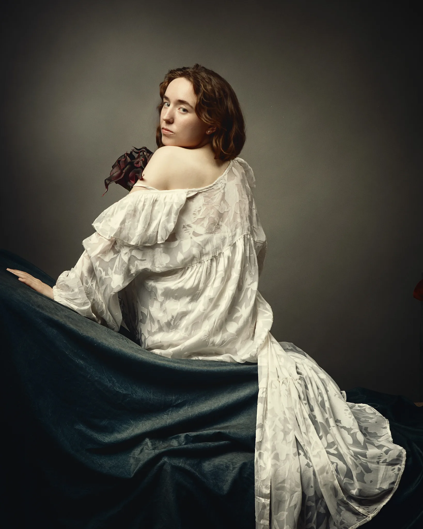 A woman with shoulder-length brown hair looks over her shoulder. She wears a white draped garment and holds a dark blue fabric. A dark red flower rests on her shoulder.