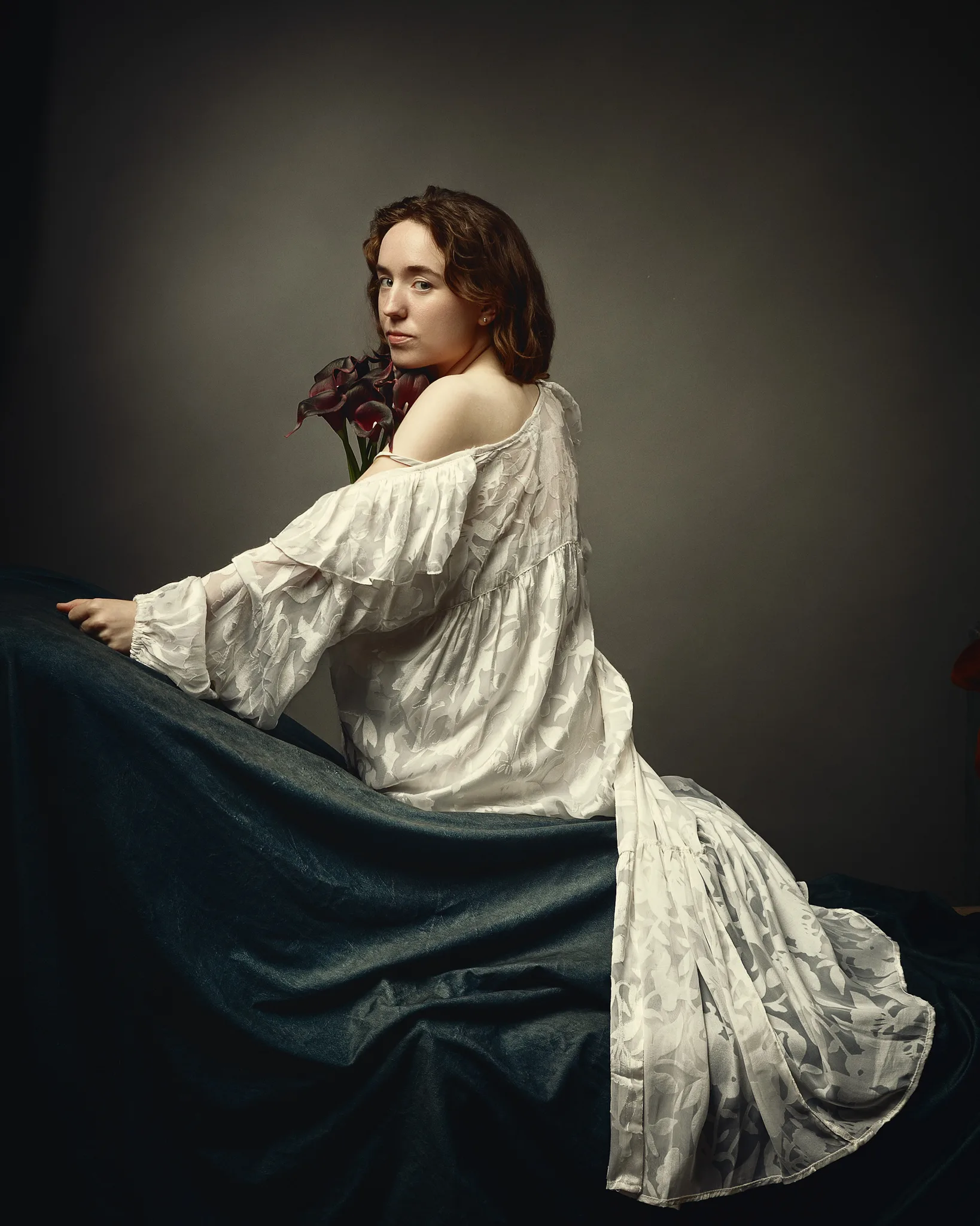 A young woman in a white draped garment holds dark red flowers, seated against a muted background, looking over her shoulder.