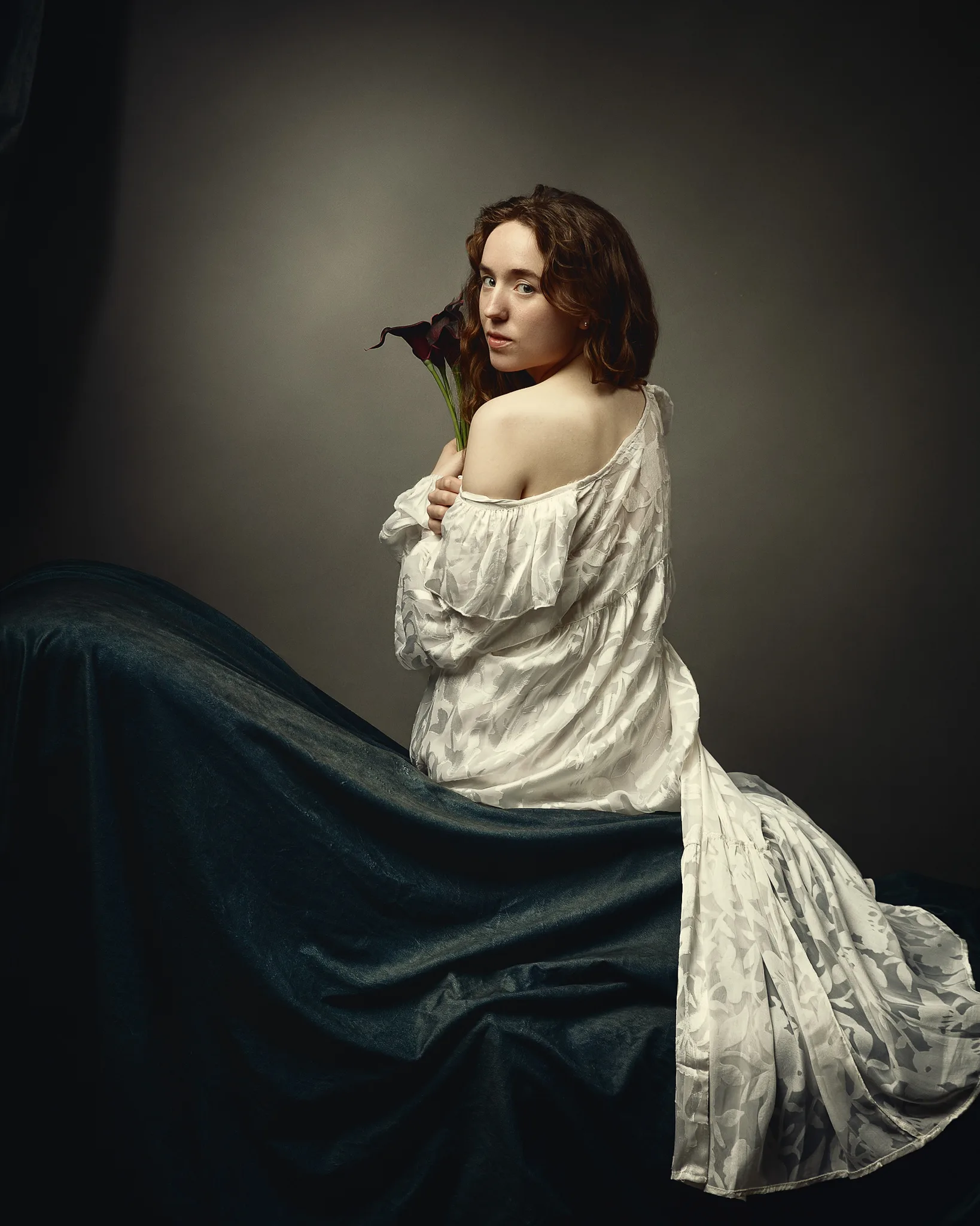 A woman with shoulder-length brown hair sits facing away, holding a dark red calla lily. She wears a white off-shoulder blouse and a dark teal fabric drapes across her lap.