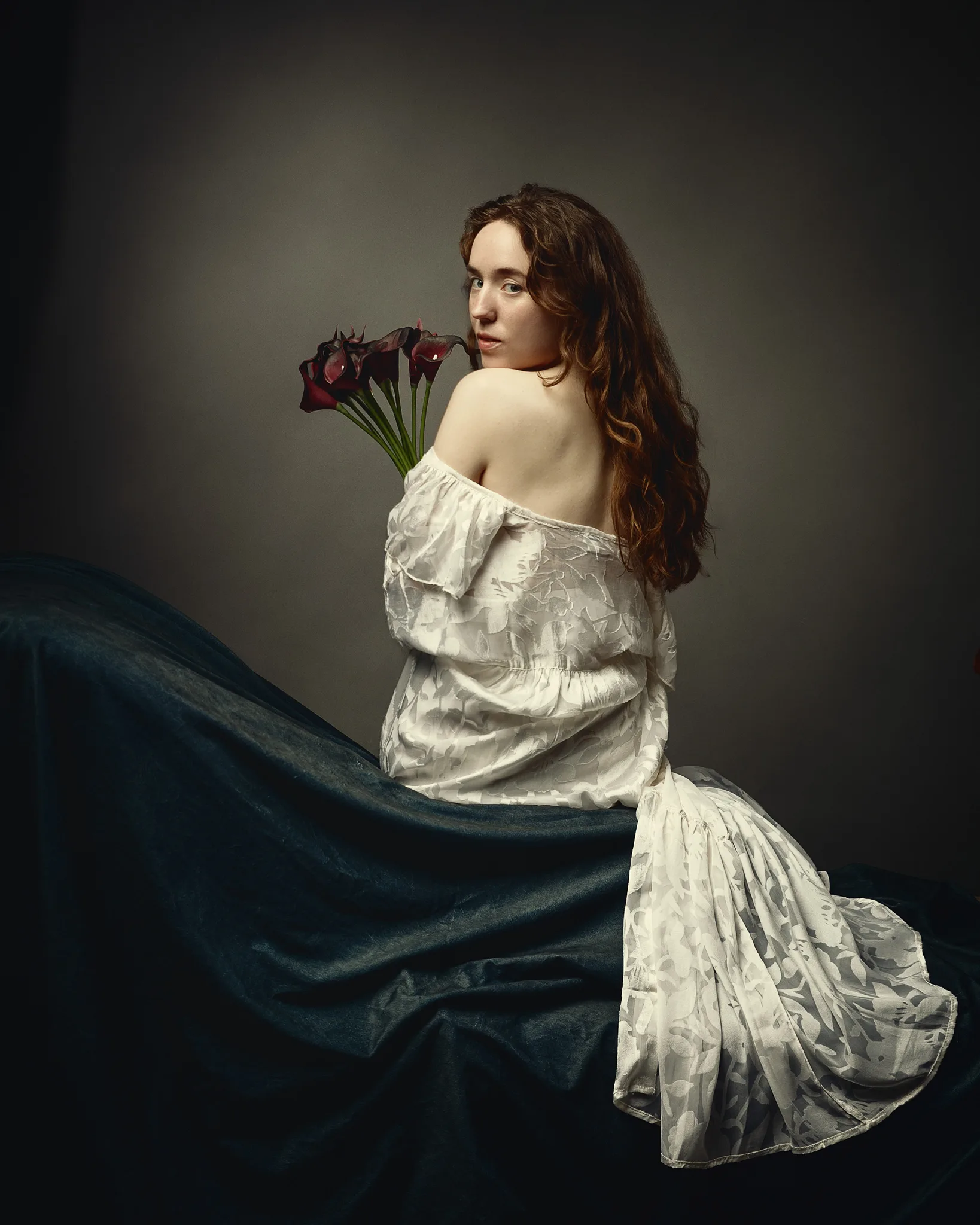 A woman with long reddish-brown hair sits holding dark red calla lilies, wearing an off-shoulder cream dress against a neutral background.
