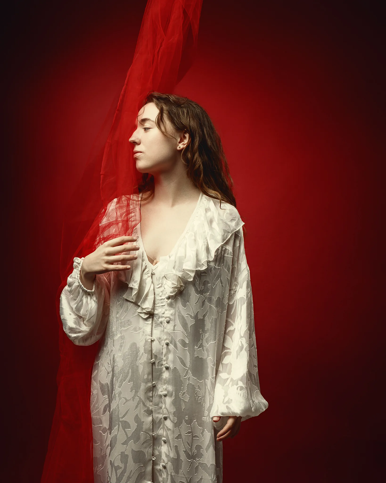 A woman in a pale patterned robe stands in profile against a red background, holding red fabric.