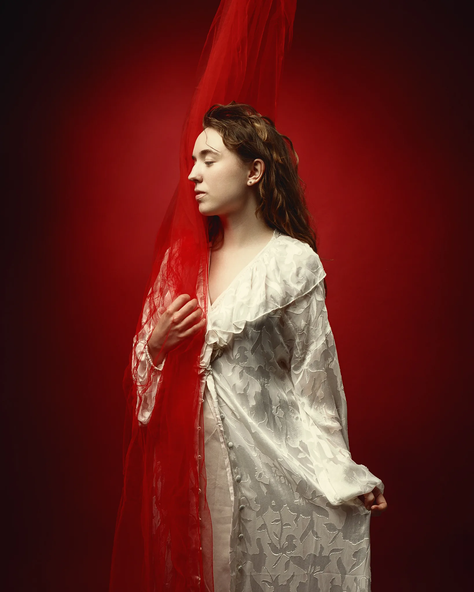 A woman in profile wearing a cream embroidered dress holds a flowing red veil against a deep red background.