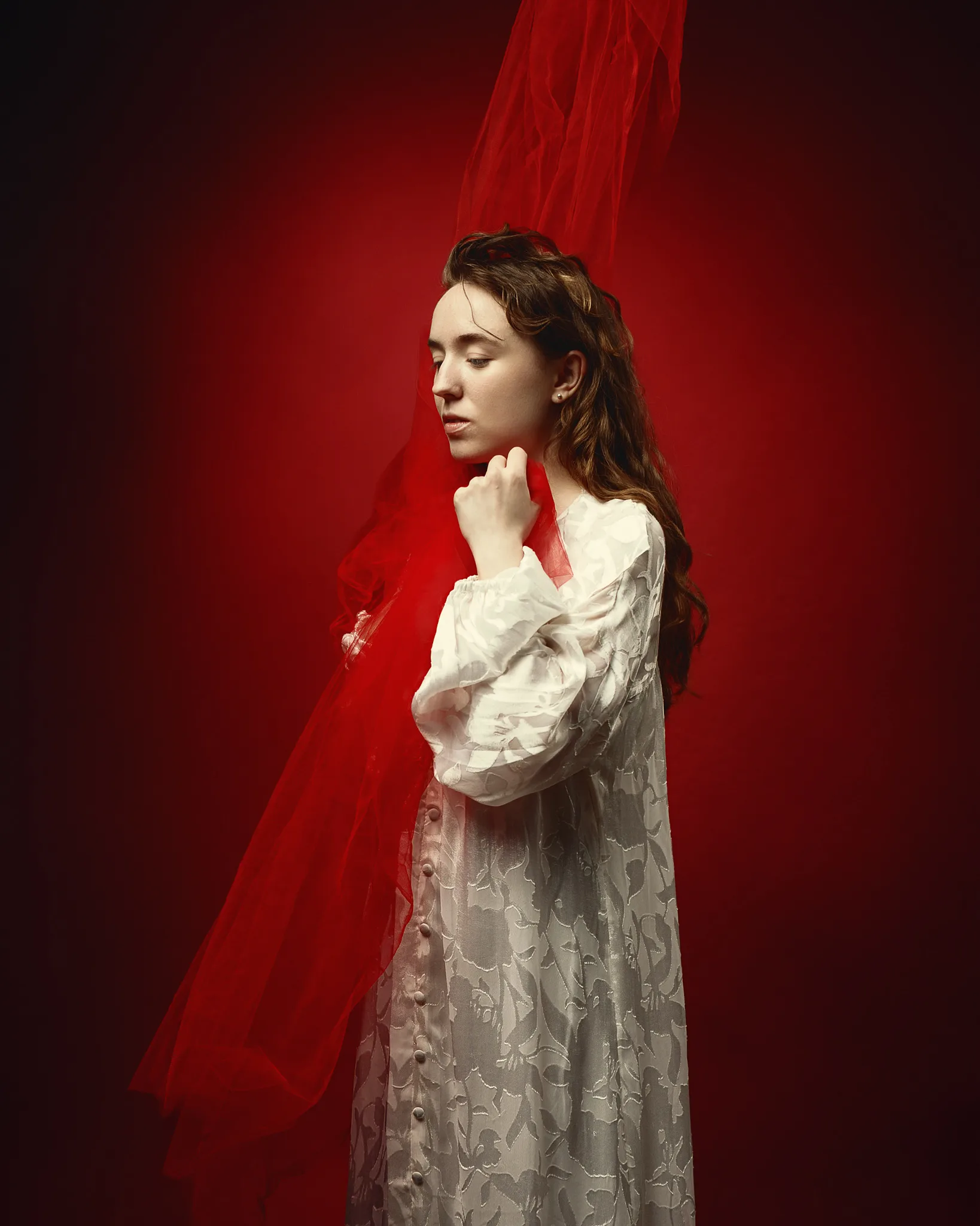 A woman in a white embroidered dress and white blouse stands in profile against a vivid red background, with red fabric draped behind her.