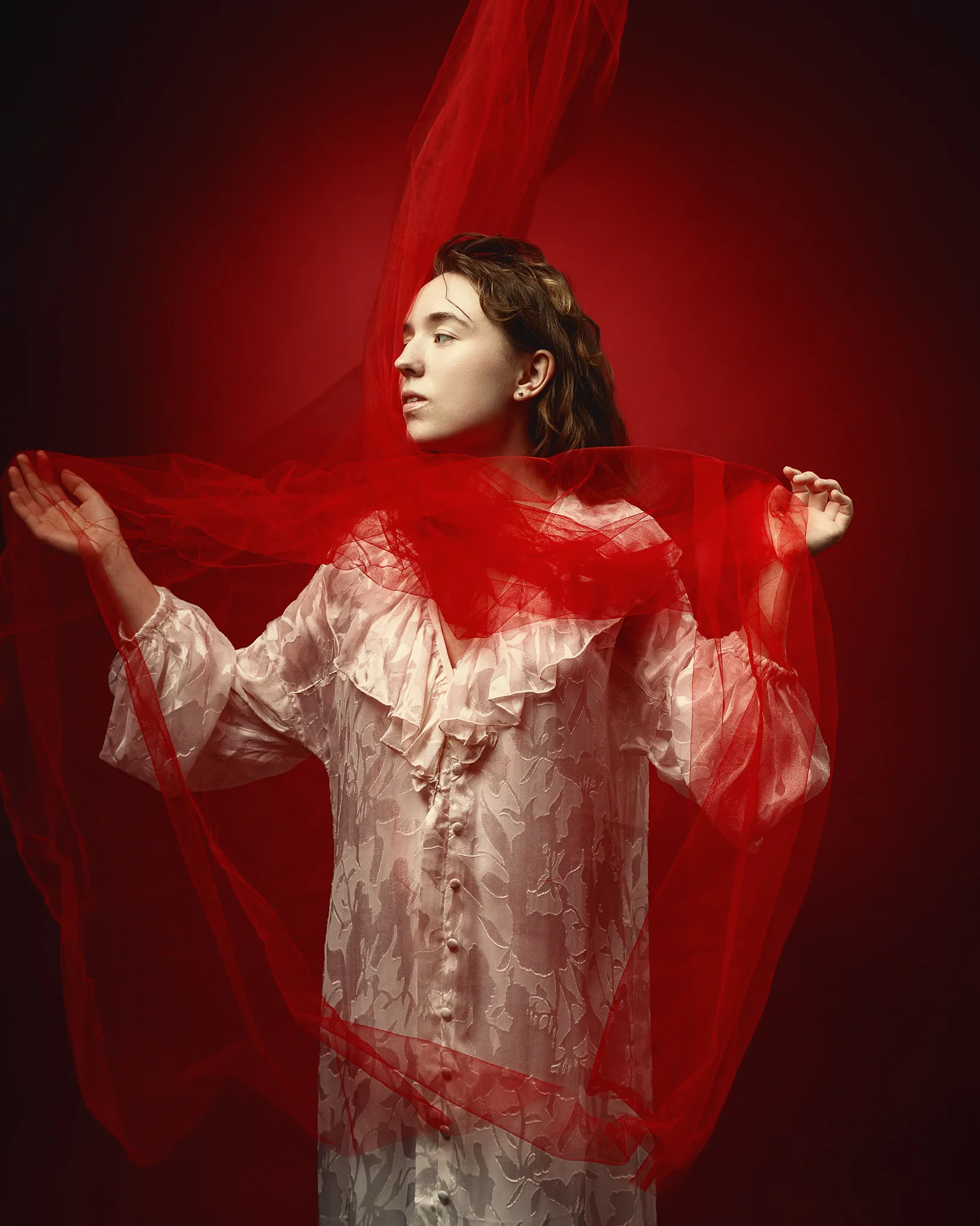 A woman in an ornate cream dress with ruffled collar stands against a red background, arms outstretched holding flowing red tulle fabric.