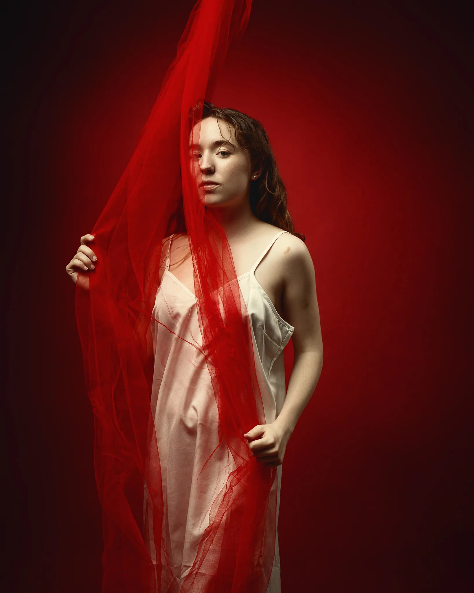 A woman in a pale slip dress holds a sheer red fabric against a deep red background, looking downward pensively.
