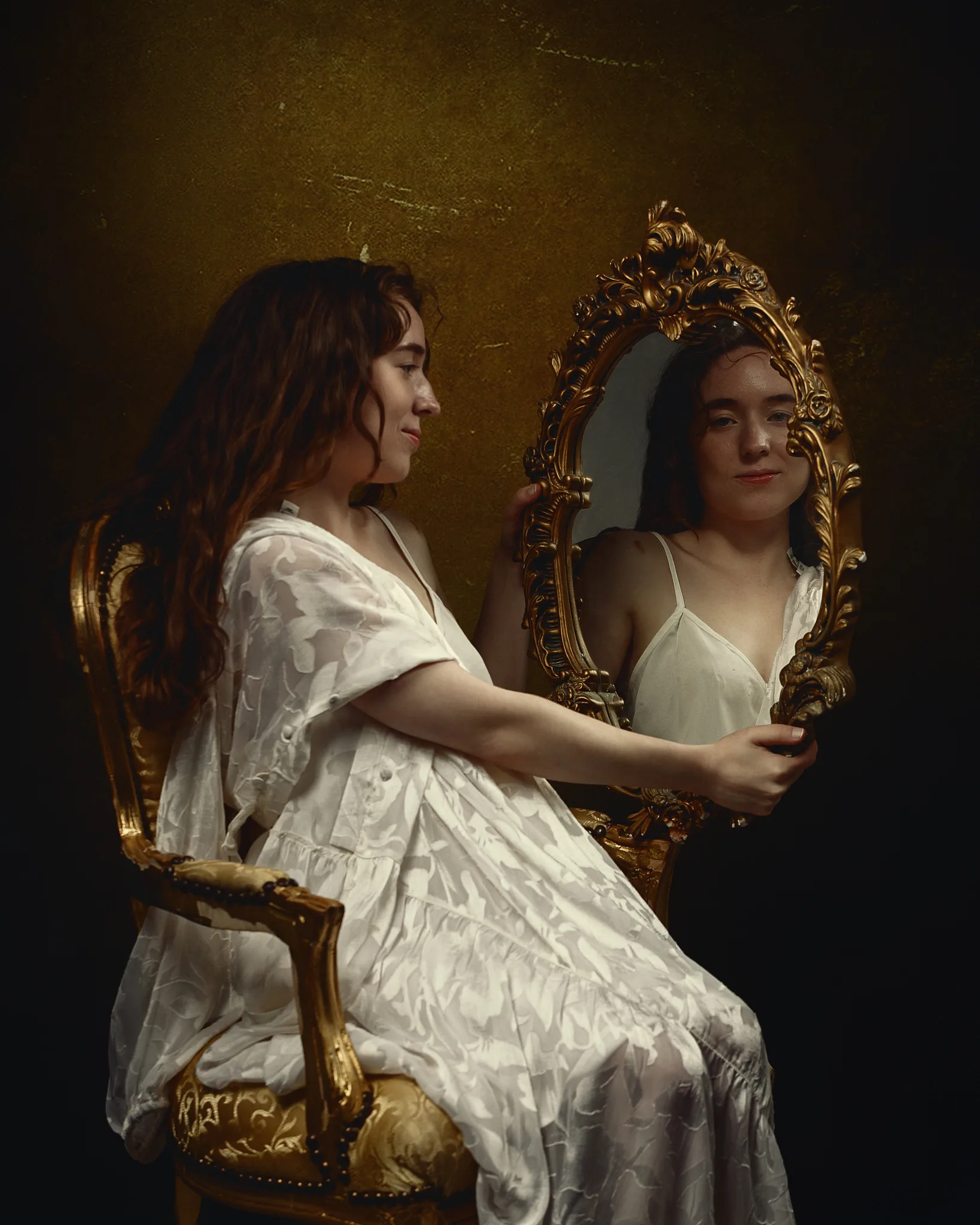 Woman in white dress holding ornate gold mirror, seated on gold chair against dark gold background.