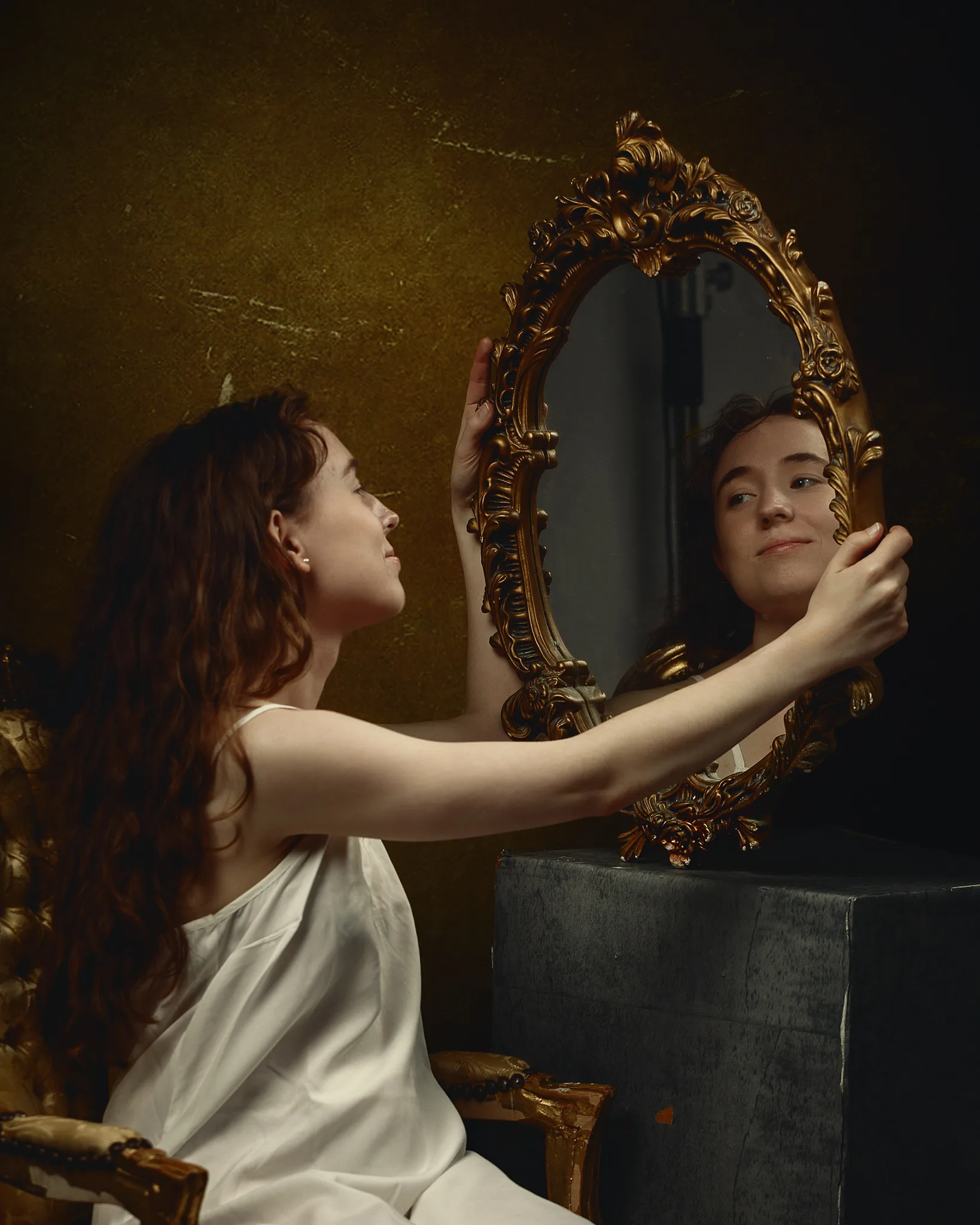 A woman in a white garment gazes into an ornate gold-framed mirror against a textured gold wall.