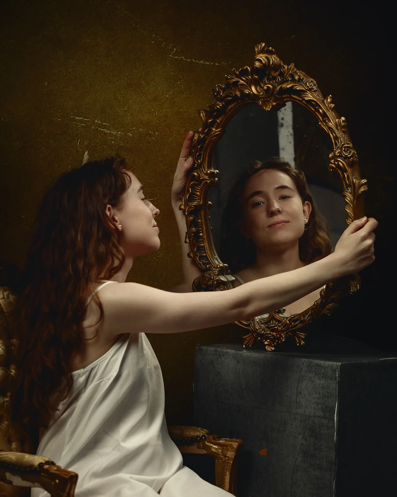 A young woman in a white garment gazes at her reflection in an ornate gold-framed mirror against a textured gold wall.