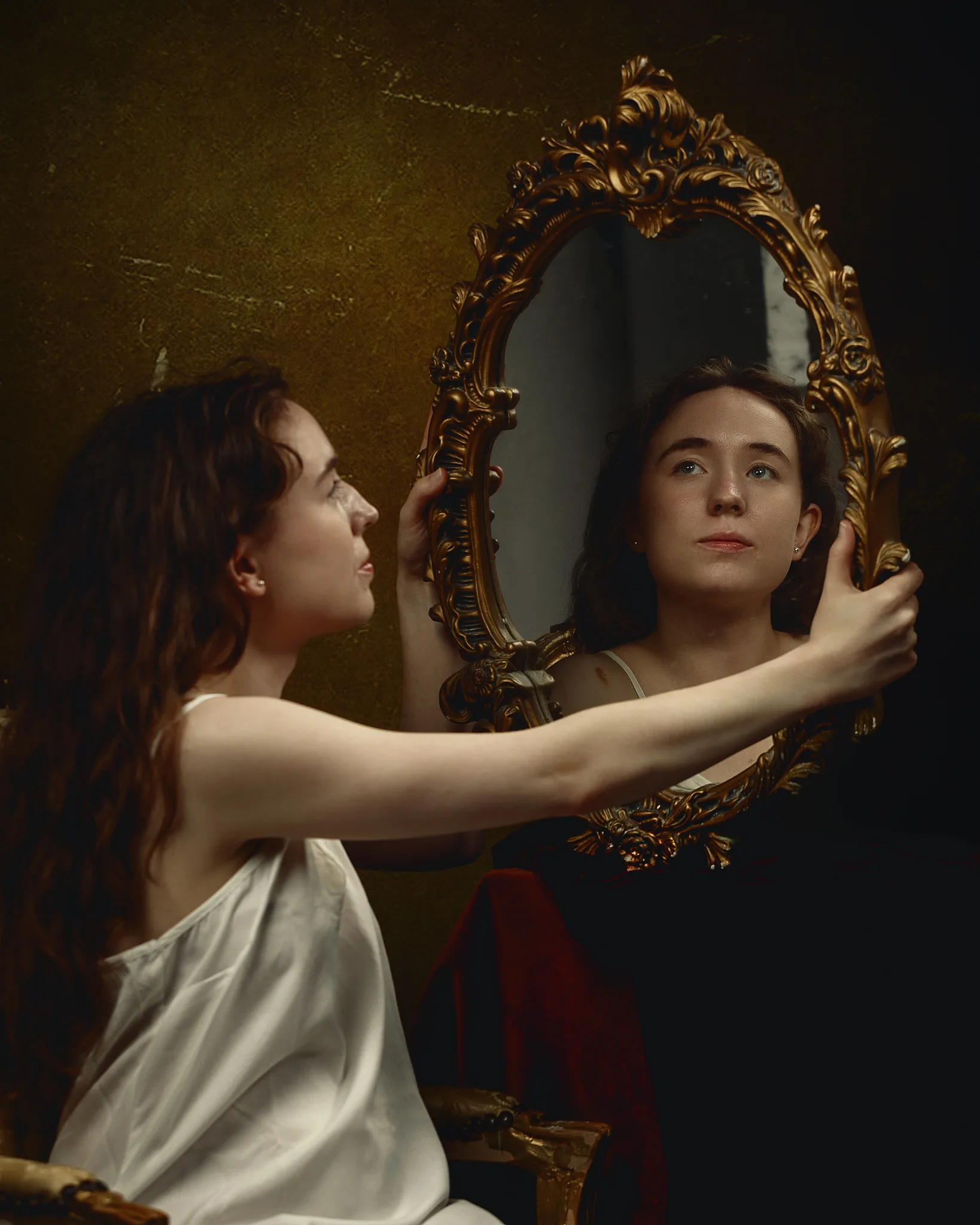 A woman in a white draped garment holds an ornate gold-framed mirror, gazing at her reflection against a dark background.